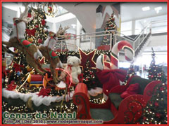 Nesta decora&ccedil;&atilde;o renas puxam um tren&oacute; vermelho do papai noel cercado por pinheiros de natal com luzes cintilantes